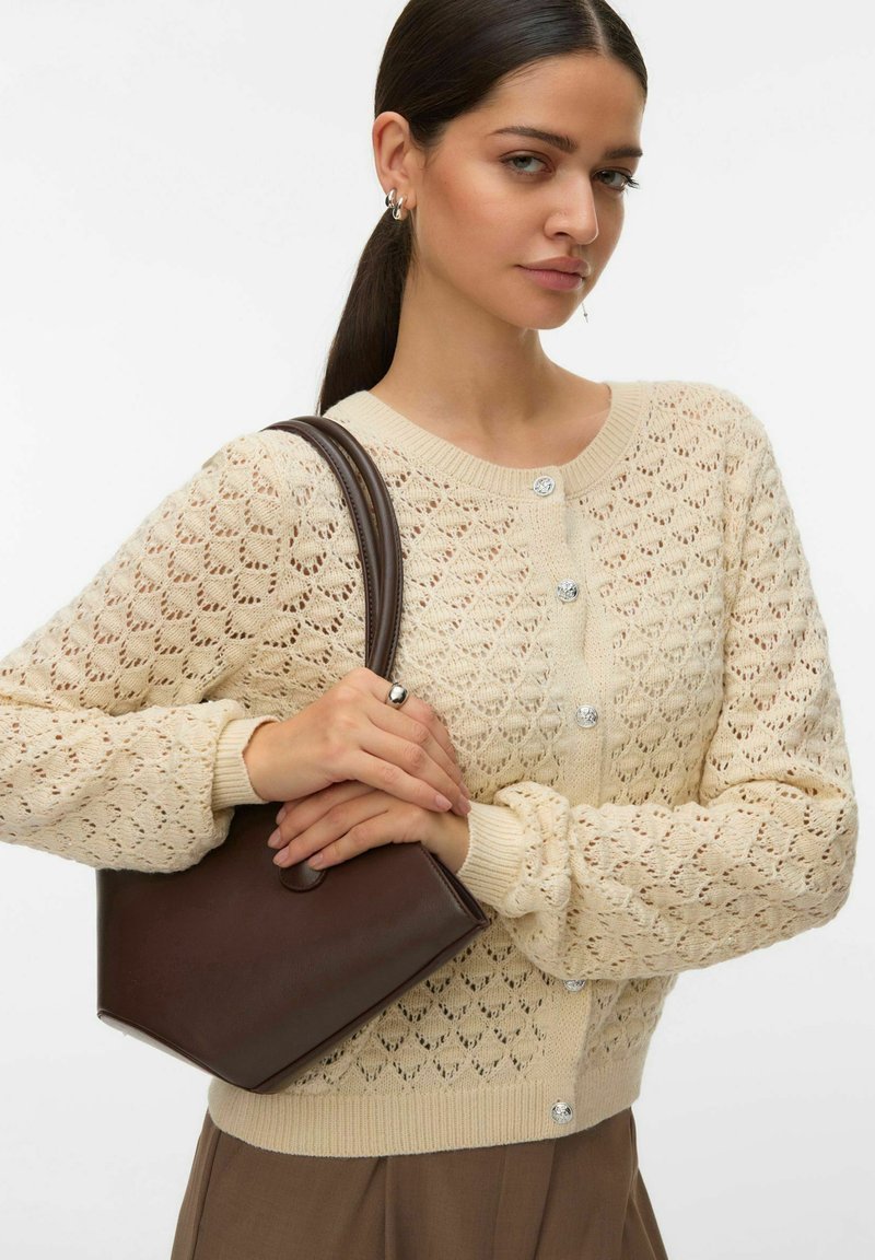 Cardigan en tricot beige avec motif ajouré, encolure ronde et boutons décoratifs. Tenant un sac à main minimaliste marron. Pantalon marron avec une texture subtile.