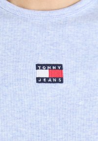 T-shirt azzurro chiaro a righe con un piccolo logo rettangolare ricamato che mostra la scritta "TOMMY" in bianco sopra un blocco rosso e "JEANS" in bianco sotto di esso.