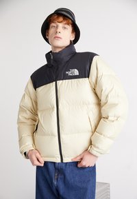 Pufferjack in crème en zwart, geïsoleerd met een gestikt ontwerp. Beschikt over een hoge kraag, voorrits en logo op de borst. Gecombineerd met denim.