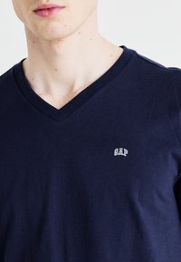 Tumed sinise V-kaelusega T-särk, mis on valmistatud puuvillast, vasakul küljel on väike valge GAP logo. Kanga tekstuur on sile ja pehme.