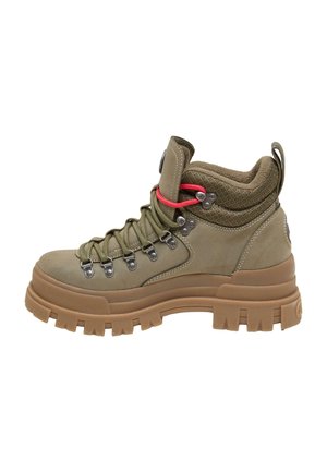 Personne portant un pantalon en cuir noir et des chaussures de randonnée épaisses couleur beige, assise sur des marches en béton à l'extérieur.