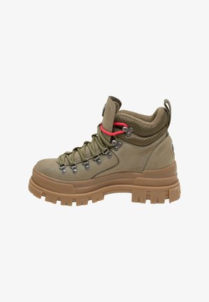 Personne portant un pantalon en cuir noir et des chaussures de randonnée épaisses couleur beige, assise sur des marches en béton à l'extérieur.