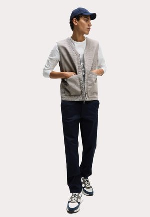 Gilet en velours côtelé gris clair avec fermeture éclair à l'avant, deux poches avant, porté sur une chemise blanche à manches longues, associé à un pantalon sombre et des baskets.