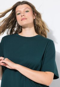 Mladá žena se světle hnědými vlnitými vlasy, zeleným očním linkem, velkými kruhovými náušnicemi a tmavě zeleným tričkem, pózuje s rukama blízko hrudníku.