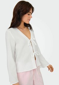 Blouse blanche texturée avec un col en V et des fermetures à lien à l'avant. Présente des manches longues et une coupe décontractée. Associée à un pantalon rayé rose clair.