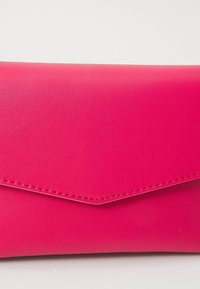 Jasně růžová kožená peněženka ve stylu obálky s viditelným prošíváním podél okraje klopy na bílém pozadí.