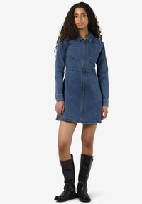 Denim skjorte kjole med lange ermer, to brystlommer, glidelås foran og en figurnær silhuett. Brukt med knelange svarte lærstøvler.