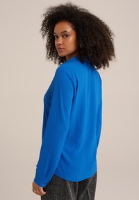 Blusa blu a maniche lunghe realizzata in un tessuto testurizzato. Presenta un colletto, polsini abbottonati e un orlo curvo. Indossata con pantaloni grigi a fantasia.