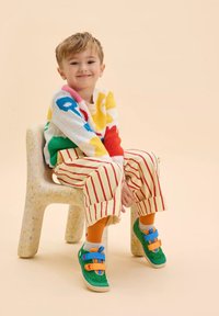 Maglione lavorato a maglia colorato con motivi diversi, pantaloni a righe e sneakers verde con velcro, decorate con accenti arancioni e blu, seduto su una sedia maculata.