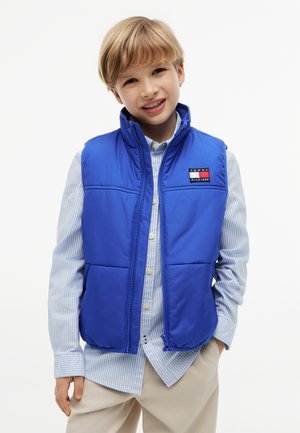 Blonder Junge lächelt, trägt eine blaue Steppweste über einem hellblau gestreiften Hemd mit Knöpfen und beigen Hose, steht mit einer Hand in der Tasche.