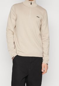 Pull beige zippé avec col et poignets côtelés, comportant un petit logo noir "BOSS" sur le côté gauche de la poitrine. Tissu en maille douce, coupe ajustée.