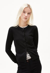 Svart cardigan i mjukt tyg med rund halsringning och knäppning. Bärs över en grå topp, har långa ärmar och en avslappnad passform.