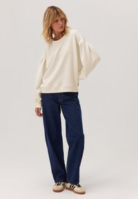 Kobieta w oversize'owej kremowej bluzie, luźnych niebieskich dżinsach i beżowych sneakersach z czarnymi paskami, stojąca na tle jasnego, jednolitego tła.