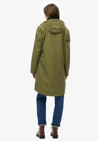 Chaqueta impermeable verde oliva con capucha, cierre frontal con cremallera y puños ajustables, combinada con jeans azules y botas marrones con cordones.