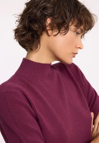 Bordowy sweter dzianinowy z prążkowanym wysokim kołnierzem, miękką teksturą, dopasowanym krojem i długimi rękawami. Zbliżenie na kołnierz i ramię.