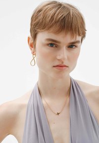 Collar de metal dorado con un pequeño colgante de cruz, acompañado de un pendiente asimétrico dorado. La modelo lleva un top halter de color púrpura claro.