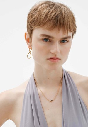 Collar de metal dorado con un pequeño colgante de cruz, acompañado de un pendiente asimétrico dorado. La modelo lleva un top halter de color púrpura claro.