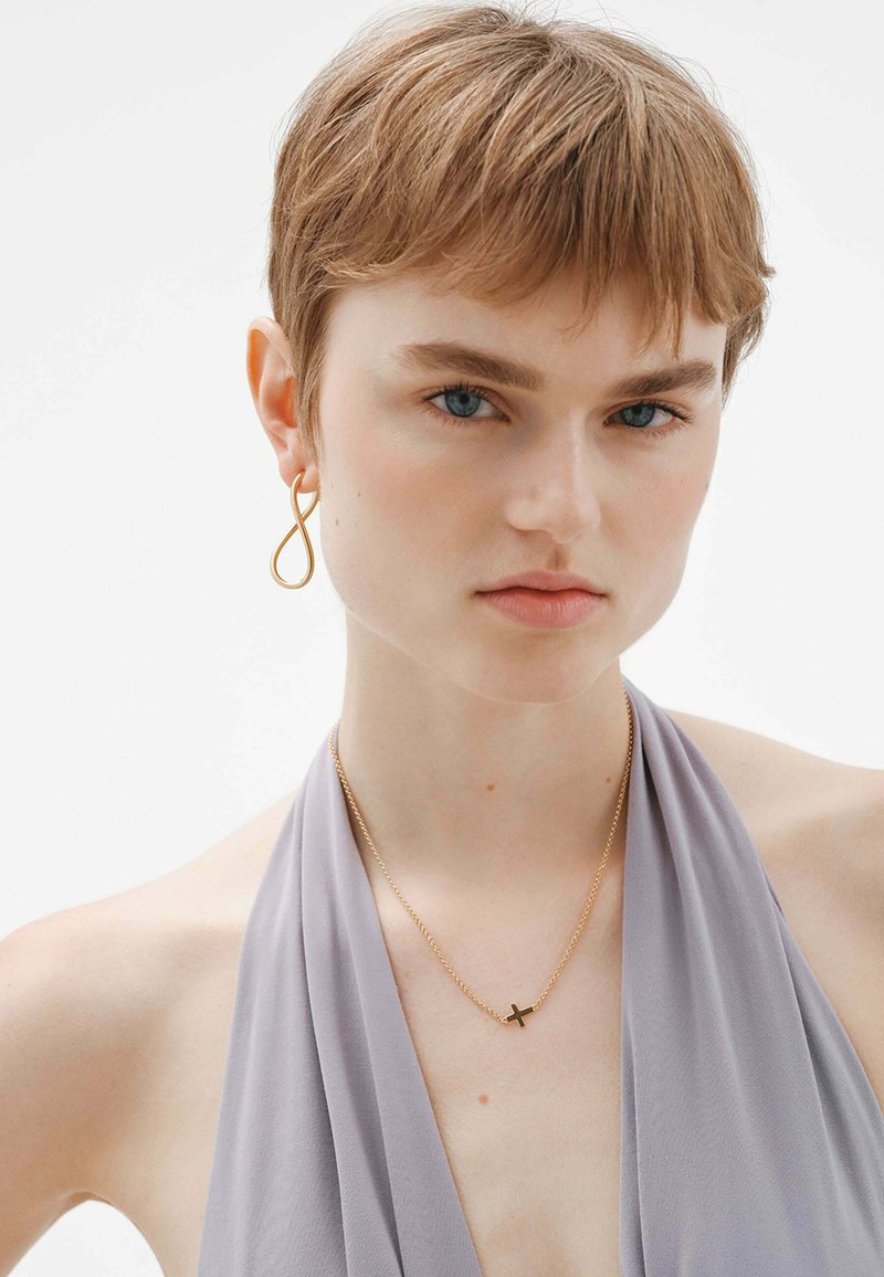 Collar de metal dorado con un pequeño colgante de cruz, acompañado de un pendiente asimétrico dorado. La modelo lleva un top halter de color púrpura claro.
