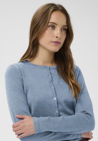 Gaiši zila garo piedurkņu kardigāna jaka, kas izgatavota no mīkstas materiāla, ar apaļu kakla izgriezumu, pogu aizdari priekšpusē un ribotu tekstūru.