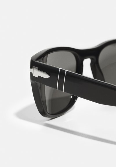 Occhiali da sole con montatura in plastica nera spessa, lenti grigie scure e un accento di design metallico argento sulla stanghetta.