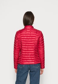 Kvinde iført en tætsiddende rød quilted jakke med albue-patcher og blå jeans, stående med ryggen mod kameraet foran en ensfarvet baggrund.