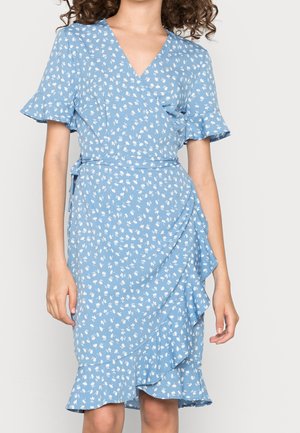Vrouw draagt een lichtblauwe wikkeljurk met een kleine witte bloemenprint, korte geplooide mouwen en ruches langs de zoom.