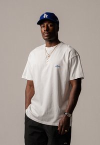 T-shirt en coton blanc avec le texte brodé bleu "ELVIS". Associé à un short noir et une casquette de baseball bleue de LA. Accents en bijoux argentés et dorés.
