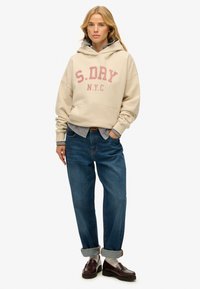 Sudadera beige con capucha y estampado "S.DAY N.Y.C.", sobre una camisa de rayas, combinada con jeans de mezclilla y mocasines oscuros.