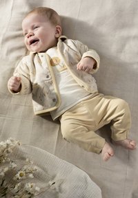 Crème-kleurige babyoutfit met een patroonjas met zakken, een grafische t-shirt en beige broek, geplaatst op een gestructureerd oppervlak.