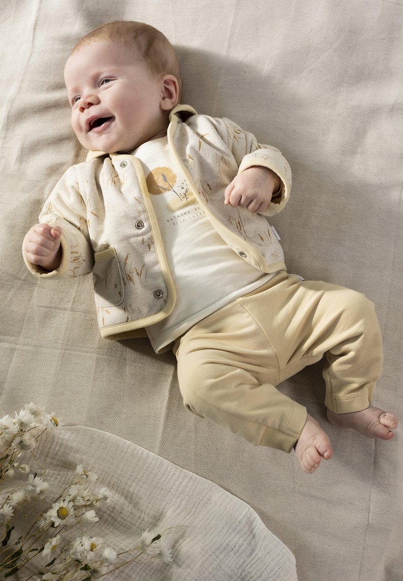 Crème-kleurige babyoutfit met een patroonjas met zakken, een grafische t-shirt en beige broek, geplaatst op een gestructureerd oppervlak.