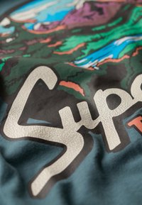 Camiseta de algodón con un colorido gráfico al aire libre que incluye árboles, montañas y un río. Logo "Super" texturizado y prominente en impresión blanca agrietada.