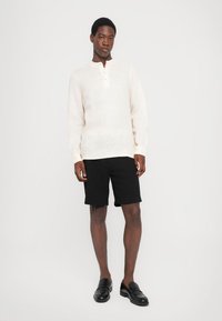 Ung mann står iført en hvit langermet henley-skjorte, svarte knelange shorts og svarte lærloafers foran en hvit bakgrunn.