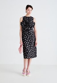 Black dress with floral pattern, oversized bow at neckline, knee-length. Model holds a pink purse; wears pink platform shoes with floral accents.