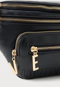 Schwarze synthetische Lederhandtasche mit mehreren goldenen Reißverschlüssen, goldenen Buchstabenanhängern "V" und "E" sowie geprägtem Text "LOVE ME" auf der Vorderseite.