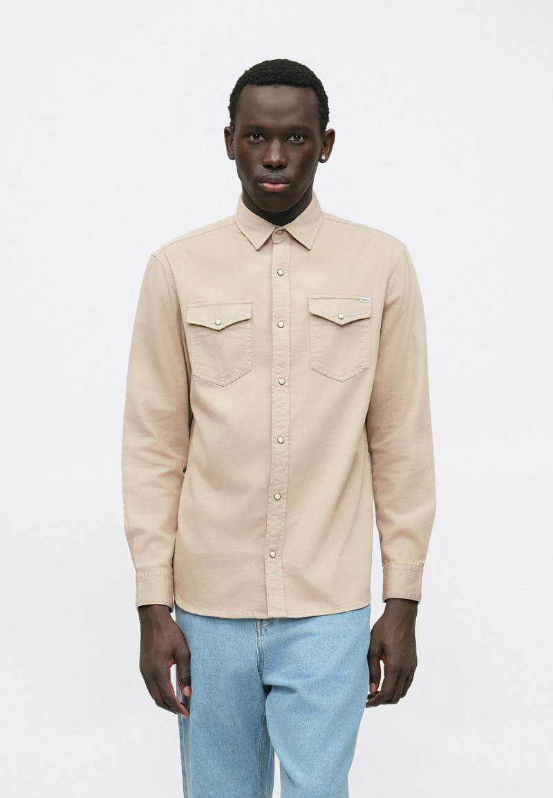 Mlad moški s kratkimi kodrastimi lasmi, oblečen v bež srajco na gumb z dvema žepoma na prsih in svetlo modre kavbojke, stoji pred enobarvnim ozadjem.