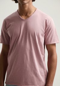Camiseta de manga corta y escote en V en suave tela rosa. La textura parece ligera y ligeramente arrugada, con un corte relajado.