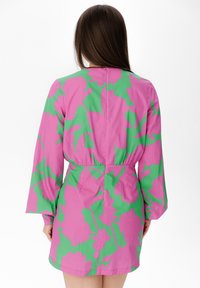 Woman with long brown hair wears a long-sleeve, short dress with a pink and green large floral pattern, viewed from the back.