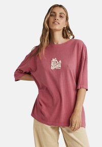 Oversized pink T-shirt with short sleeves and a crew neck, featuring a white, stylized "Billabong" logo on the front. Casual cotton fabric.