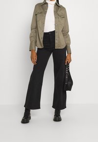 Olivgrön knapp-up skjorta, vit ribbad polotröja, högmidjade svarta vida byxor, svarta kängor, och en svart handväska med metalldetaljer.