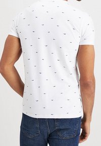 Camisa polo blanca de manga corta con pequeños patrones azules, hecha de un tejido suave, con cuello redondo y un diseño ajustado.