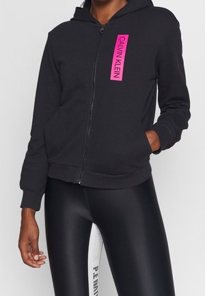 Sudadera negra con cremallera y parche rosa con el logo de Calvin Klein, usada con leggings negros y blancos con texto en el panel blanco.