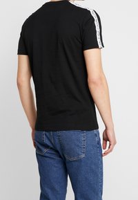 Muž v černém tričku s krátkým rukávem, na kterém jsou bílé pruhy s logem Calvin Klein Jeans na ramenou, doplněném modrými džíny, stojící zády.