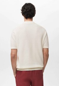Cream-colored short-sleeve polo shirt with a textured knit pattern. Features a flat collar and ribbed hems. Worn with dark red pants.