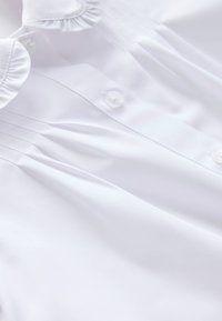 Weiße Bluse mit einem Rüschenkragen, einer Knopfleiste und weichem, glattem Stoff. Dezente Falten verleihen dem Design Struktur.