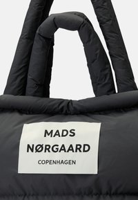 Bolsa tote acolchoada preta com alças grossas e suaves. Apresenta um rótulo branco com texto preto a dizer "MADS NØRGAARD COPENHAGUE." Textura suave.