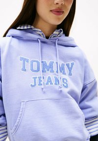 Felpa leggera viola con "TOMMY JEANS" ricamato in blu. Presenta una tasca frontale e, sotto, una camicia colorata blu e bianca a righe con colletto.