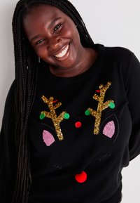 Schwarzer Pullover mit einem Rentierdesign, das goldene Pailletten-Hörner, bunte Filzornamente und pinke Akzente im Gesicht hat.