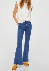 Haut blanc avec des manches volantées et des détails en dentelle, associé à un jean bleu évasé et des sandales jaunes, affichant un style décontracté.