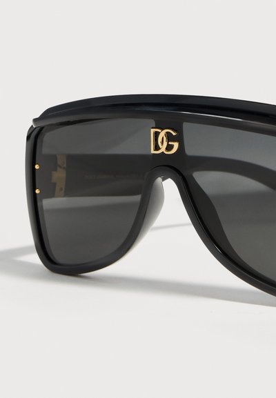 Oversized μαύρα γυαλιά ηλίου σε σχήμα ασπίδας με σκουρόχρωνους φακούς και χρυσό λογότυπο DG στο κέντρο πάνω από τη γέφυρα της μύτης σε λευκό φόντο.