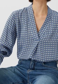 Blå mönstrad blus med en inslagning design, prydd med geometriska former och en djup V-ringning, ihopparad med högmidjade denimjeans.